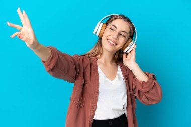 Genç beyaz kadın mavi arka planda izole edilmiş müzik dinliyor ve şarkı söylüyor.