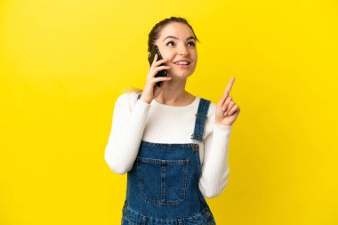 İzole edilmiş sarı arka plan üzerinde cep telefonu kullanan genç bir kadın harika bir fikre işaret ediyor.