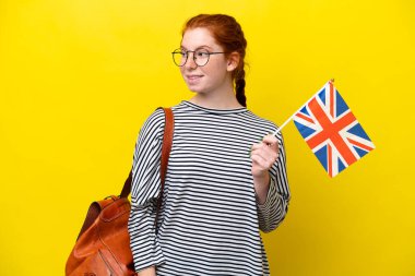 Sarı arka planda izole edilmiş Birleşik Krallık bayrağını tutan genç İspanyol kadın yana bakıyor ve gülümsüyor.