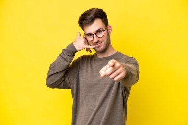 Young caucasian man isolated on yellow background making phone gesture and pointing front