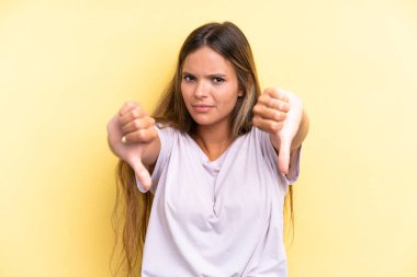 Sarı arka planda izole edilmiş genç beyaz kadın baş parmağını iki eliyle aşağı gösteriyor.