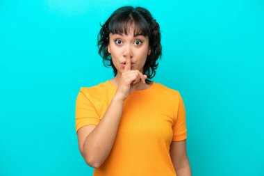 Arjantinli genç bir kadın, mavi arka planda izole edilmiş. Parmağını ağzına koyduğunda sessizlik işareti gösteriyor.