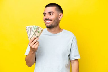 Kafkasyalı genç adam, sarı arka planda izole edilmiş bir şekilde bir sürü para alıyor. Yana bakıyor ve gülümsüyor.