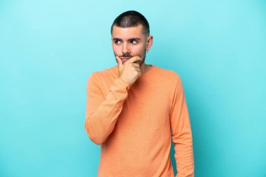 Kafkasyalı genç adam mavi arka planda izole edilmiş. Şüpheleri var ve kafası karışmış bir yüz ifadesi var.