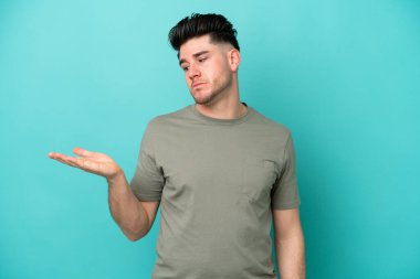 Mavi arka planda izole edilmiş genç beyaz adam, kopyalama alanını şüphe ile tutuyor.
