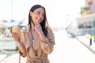 Açık havada elinde kahve tutan genç bir kadın bir fikir üretiyor ve bakıyor.