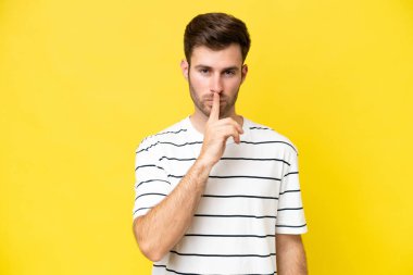 Sarı arka planda izole edilmiş genç beyaz bir adam sessizlik işareti gösteriyor. Parmağını ağzına sokuyor.