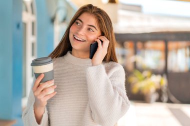 Genç beyaz bir kadın cep telefonu kullanıyor ve elinde mutlu bir ifadeyle kahve tutuyor.