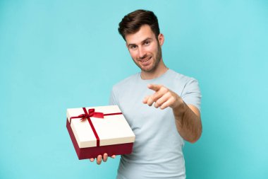 Elinde mavi arka planda izole edilmiş bir hediye tutan genç beyaz adam kendine güvenen bir ifadeyle seni işaret ediyor.