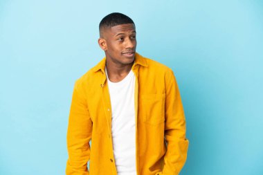 Genç latin adam mavi arka planda izole edilmiş, yana bakıyor ve gülümsüyor.