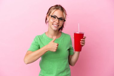 Pembe arka planda izole edilmiş serinletici bir şey tutan genç Rus kadın baş parmak işareti yapıyor.