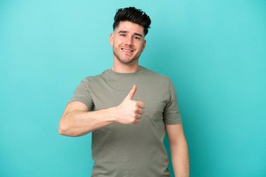 Kafkasyalı genç adam mavi arka planda izole edilmiş bir şekilde baş parmak işareti yapıyor.