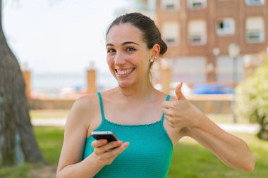 Dışarıda cep telefonu kullanan genç ve güzel bir kadın. Başparmakları havada. Çünkü iyi bir şey oldu.