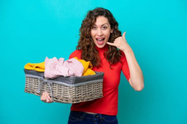 Mavi arka planda izole edilmiş çamaşır sepetini tutan beyaz bir kadın telefon jesti yapıyor. Bana geri dönüş işareti ver.