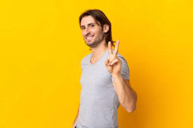 Sarı arka planda saklanmış yakışıklı genç adam gülümsüyor ve zafer işareti gösteriyor.