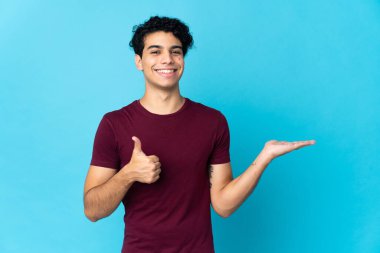 Genç Venezuelalı bir adam mavi arka planda izole edilmiş. Kopyalama alanı hayali bir şekilde avucunun içinde duruyor. Bir ilan vermek için.