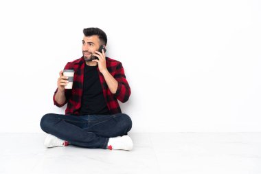 Yerde oturan yakışıklı genç bir adam elinde kahve ve bir cep telefonu tutuyor.