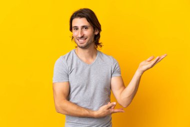 Sarı arka planda izole edilmiş genç ve yakışıklı bir adam davet edildiği için ellerini yana uzatıyor.