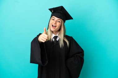 Tecrit edilmiş mavi arka planda genç üniversite mezunu. Başparmakları havada. Çünkü iyi bir şey oldu.