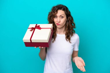 Mavi arka planda izole edilmiş bir hediye tutan beyaz bir kadın omuzlarını kaldırırken şüpheyle hareket ediyor.