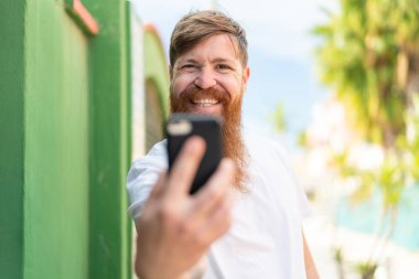 Kızıl saçlı, sakallı, dışarıda cep telefonu kullanan neşeli bir adam.