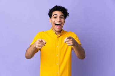 Genç Venezuelalı adam mor arka planda izole edilmiş, şaşırmış ve ön tarafı işaret ediyor.