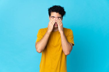 Genç Venezuelalı adam mavi arka planda izole edilmiş gözleri elleriyle kaplayan