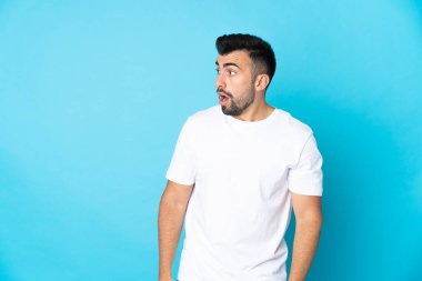 İzole edilmiş mavi arka plan üzerinde beyaz bir adam yan tarafa bakarken sürpriz jest yapıyor.