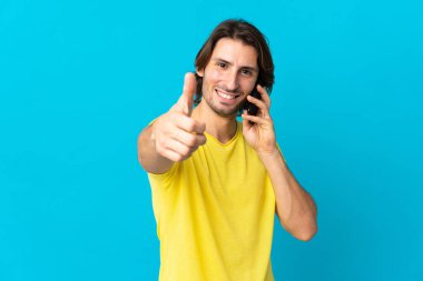 Genç ve yakışıklı bir adam mavi arka planda izole edilmiş. Başparmaklarını kaldırırken cep telefonuyla konuşuyor.