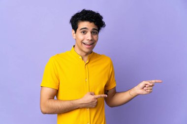 Genç Venezuelalı adam, mor arka planda izole edilmiş. Şaşırmış ve işaret ediyor.