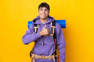 Young mountaineer man with a big backpack over isolated yellow background giving a thumbs up gesture