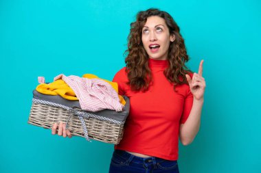 Kafkasyalı genç bir kadın mavi arka planda izole edilmiş bir çamaşır sepetini tutarken parmağıyla işaret etmeyi düşünüyor.