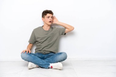 Genç Rus adam, beyaz arka planda izole bir şekilde yerde oturuyor. Kulağına elini sürerek bir şeyler dinliyor.