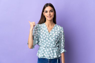 Mavi arka planda izole edilmiş genç beyaz kadın bir ürünü sunmak için kenarı işaret ediyor.