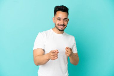 Mavi arka planda izole edilmiş genç beyaz adam şaşırmış ve ön tarafı işaret ediyor.