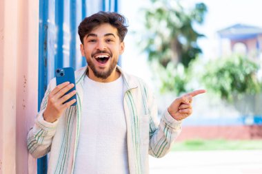 Yakışıklı bir Arap açık havada cep telefonu kullanıyor. Şaşırmış ve parmağıyla işaret ediyor.