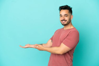 Mavi arka planda izole edilmiş genç beyaz adam, davet edilmek için ellerini yana uzatıyor.