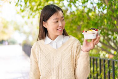 Genç Çinli bir kadın açık havada bir kase meyveyi mutlu bir ifadeyle tutuyor.