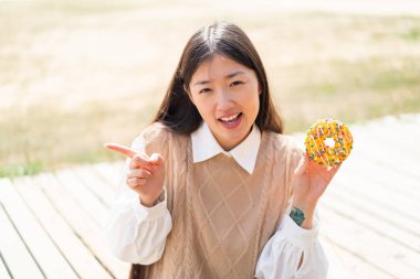 Açık havada donut tutan genç Çinli kadın şaşırdı ve parmağını yana çevirdi.