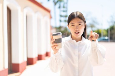 Güzel bir Çinli kadın açık havada kahve tutuyor. Şaşırmış ve ön tarafı işaret ediyor.