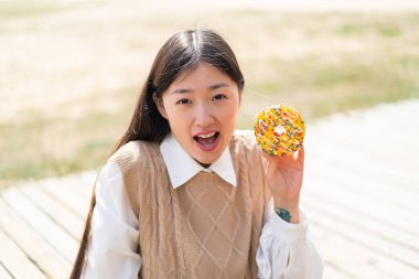 Genç Çinli bir kadın sürpriz ve şok olmuş bir yüz ifadesiyle dışarıda donut tutuyor.