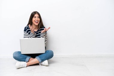 Yerde dizüstü bilgisayarlı beyaz bir kadın oturuyor. Yüzünde sürpriz bir ifade var.