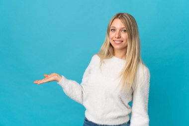 Mavi arka planda izole edilmiş genç sarışın kadın davet etmek için ellerini yana uzatıyor.