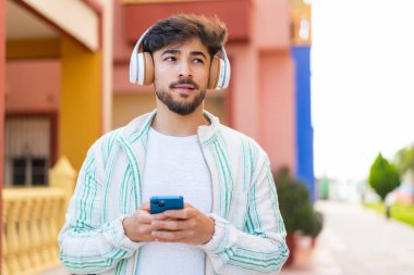 Yakışıklı bir Arap açık havada cep telefonu ile müzik dinliyor ve düşünüyor.