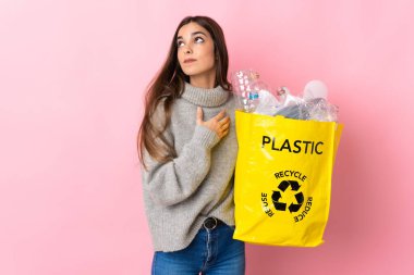 Pembe arka planda izole edilmiş plastik şişelerle dolu bir çanta taşıyan beyaz bir kadın gülümserken yukarı bakıyor.