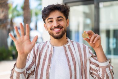 Yakışıklı bir Arap, elinde Bitcoin tutarak mutlu bir ifadeyle selamlıyor.