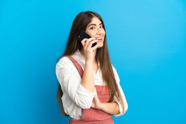 Mavi arka planda izole edilmiş genç beyaz kadın cep telefonuyla sohbet ediyor.