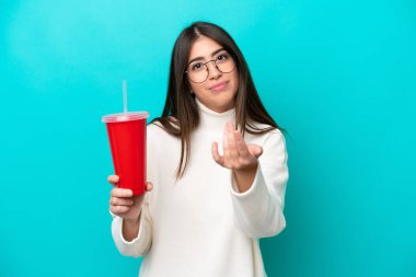 Mavi arka planda izole edilmiş soda içen genç beyaz bir kadın benimle gelmek için davet ediyor. Gelmene sevindim.