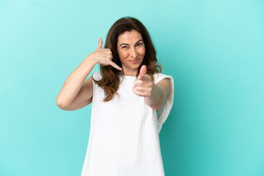 Orta yaşlı, beyaz bir kadın, mavi arka planda izole edilmiş, telefon hareketi yapıyor ve ön tarafı işaret ediyor.