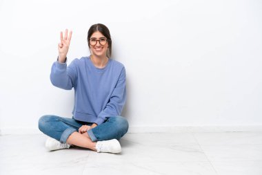 Beyaz arka planda tek başına yerde oturan beyaz bir kadın mutlu ve parmaklarıyla üçe kadar sayıyor.